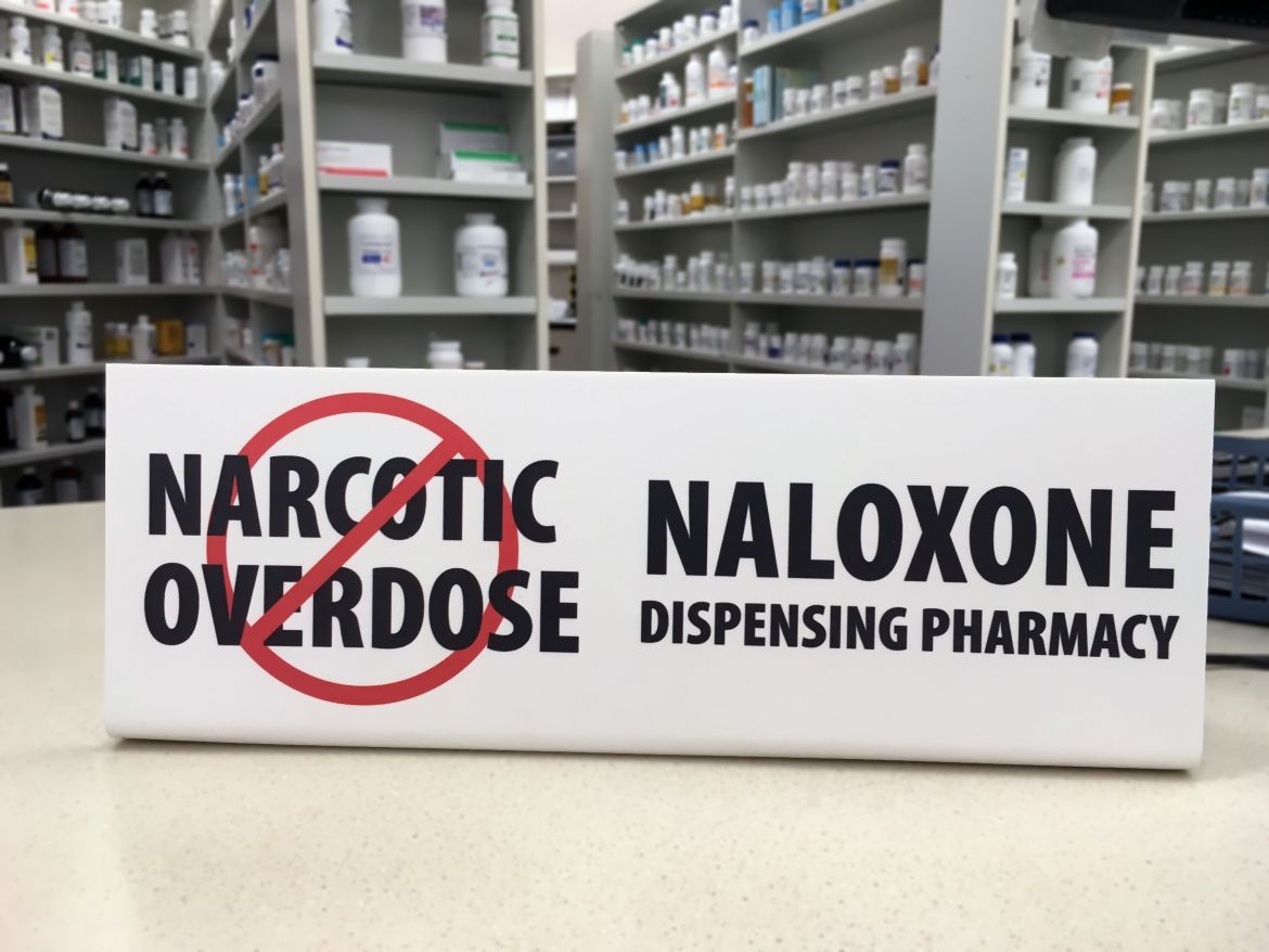 A sign for naloxone on a pharmacy counter
