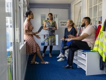 A group of teachers meet outside of a staff room. One holds a clipboard.
