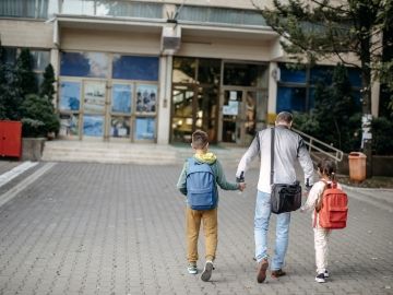 A parents walks their two children wearing backpacks towards a shool.
