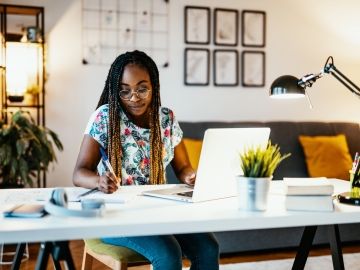 A woman sits at her desk with a laptop and takes notes.