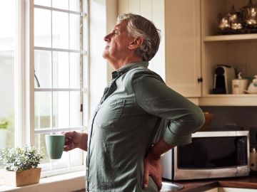 A man stands in his home with a cup of tea while holding his back in pain.