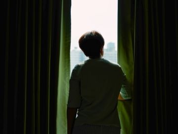 A person stands in the opening of two dark curtains, looking out the window.