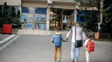 A parents walks their two children wearing backpacks towards a shool.