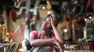 A woman worker in an apron sits inside a restaurant with her head in her hand.
