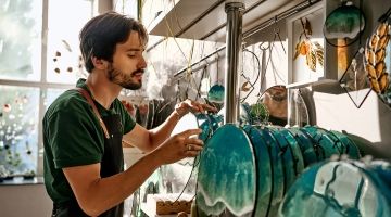 A bearded male worker in an apron handles decorative blue-teal glass discs displayed on a shelf