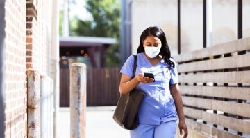 A healthcare worker in scrubs and a mask walks to work with her bag and phone in-hand.