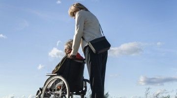A professional woman pushes an older person in a wheelchair in the outdoors