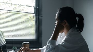 A woman working at a laptop looks out the window with her head resting on her hand