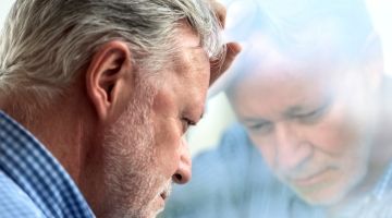 An grey-haired man leans his forehead against a window in deep contemplation
