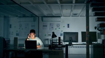 A man works at a computer in an dark, empty office late at night, with his head in his hand 