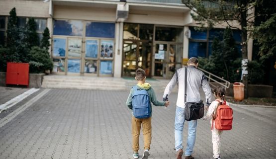 A parents walks their two children wearing backpacks towards a shool.
