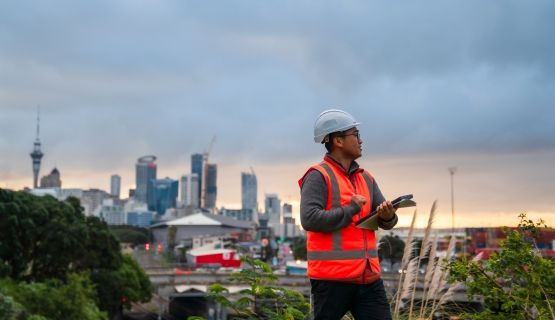 A New Zealand construction worker holding papers looking off-camera with a city skyline behind