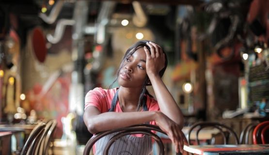 A woman worker in an apron sits inside a restaurant with her head in her hand.