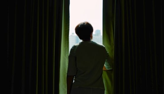 A person stands in the opening of two dark curtains, looking out the window.