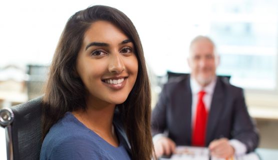 A young woman sits in front of the desk of an older man in a suit and tie, both smiling