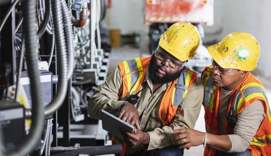 Two workers in hard hats and safety vests look at document