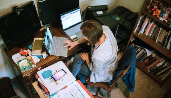 An overhead shot of a teacher, sitting in front of two laptops in a cramped corner of her home office