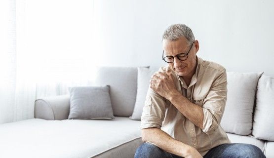 A man sitting on a couch holds his shoulder in pain