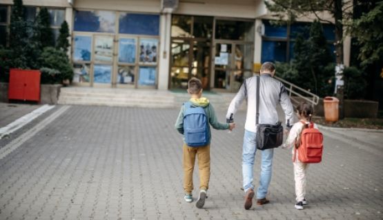 A parents walks their two children wearing backpacks towards a shool.