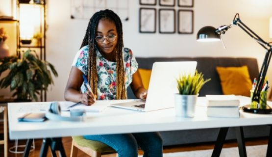 A woman sits at her desk with a laptop and takes notes.