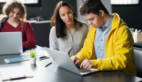 A young man working on a laptop as a female instructor guides him