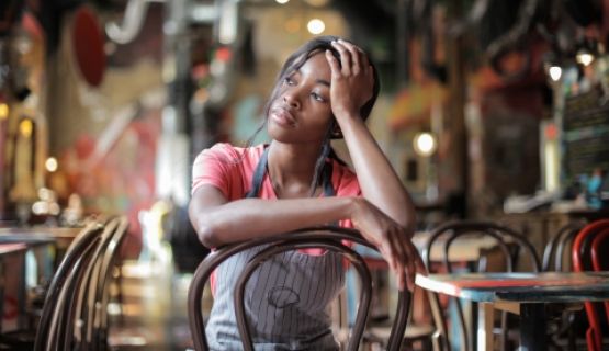 A woman worker in an apron sits inside a restaurant with her head in her hand.