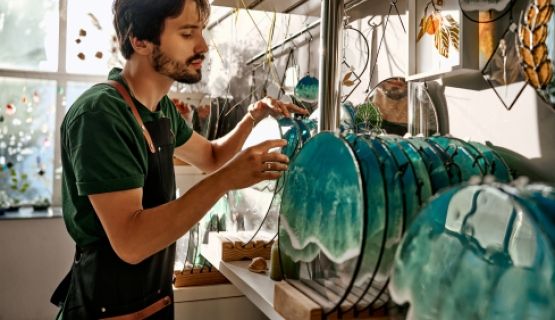 A bearded male worker in an apron handles decorative blue-teal glass discs displayed on a shelf