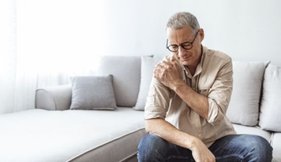 A man sitting on a couch holds his shoulder in pain